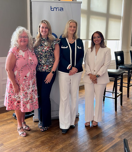 Teresa Beach-Shelow stands confidently in the foreground, wearing a black outfit. Behind her, four young women stand together in an industrial training environment, all smiling.